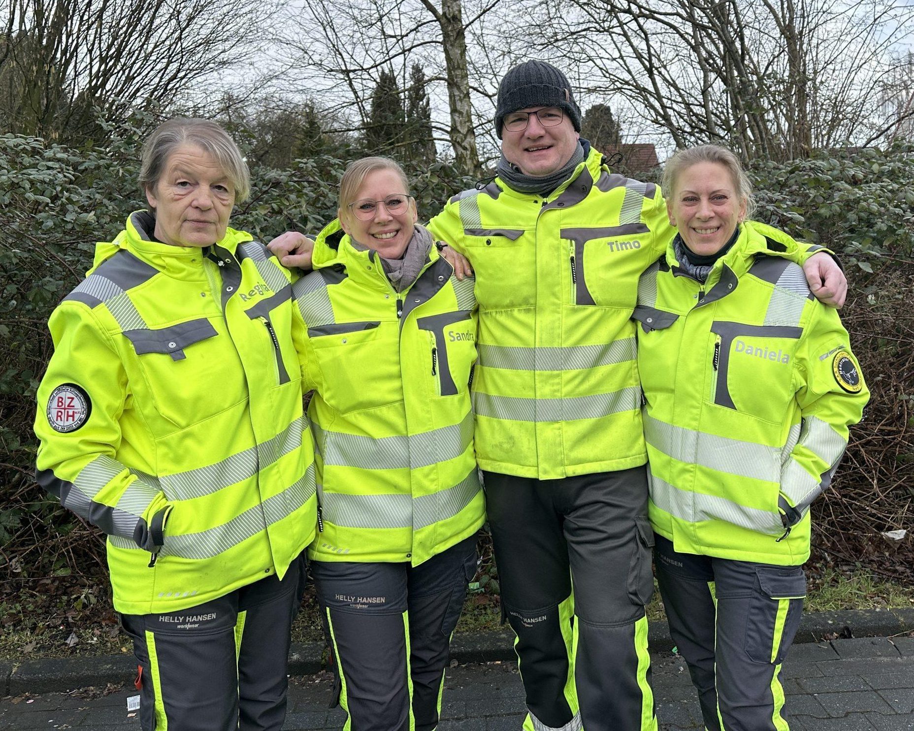 Vier Mitglieder der S.A.R. Suchhundestaffel Nord/West e.V. stehen lächelnd nebeneinander in gelber Einsatzkleidung, die Arme jeweils um die Schultern der anderen gelegt.