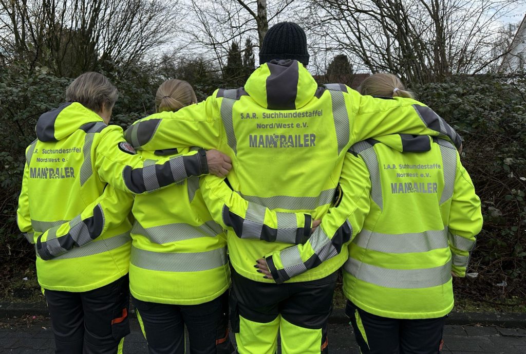 Vier Mitglieder der S.A.R. Suchhundestaffel Nord/West e.V. in gelber Einsatzkleidung mit der Aufschrift ‚MANTRAILER‘ stehen eng beisammen, mit den Armen umeinander gelegt, Rücken zur Kamera.