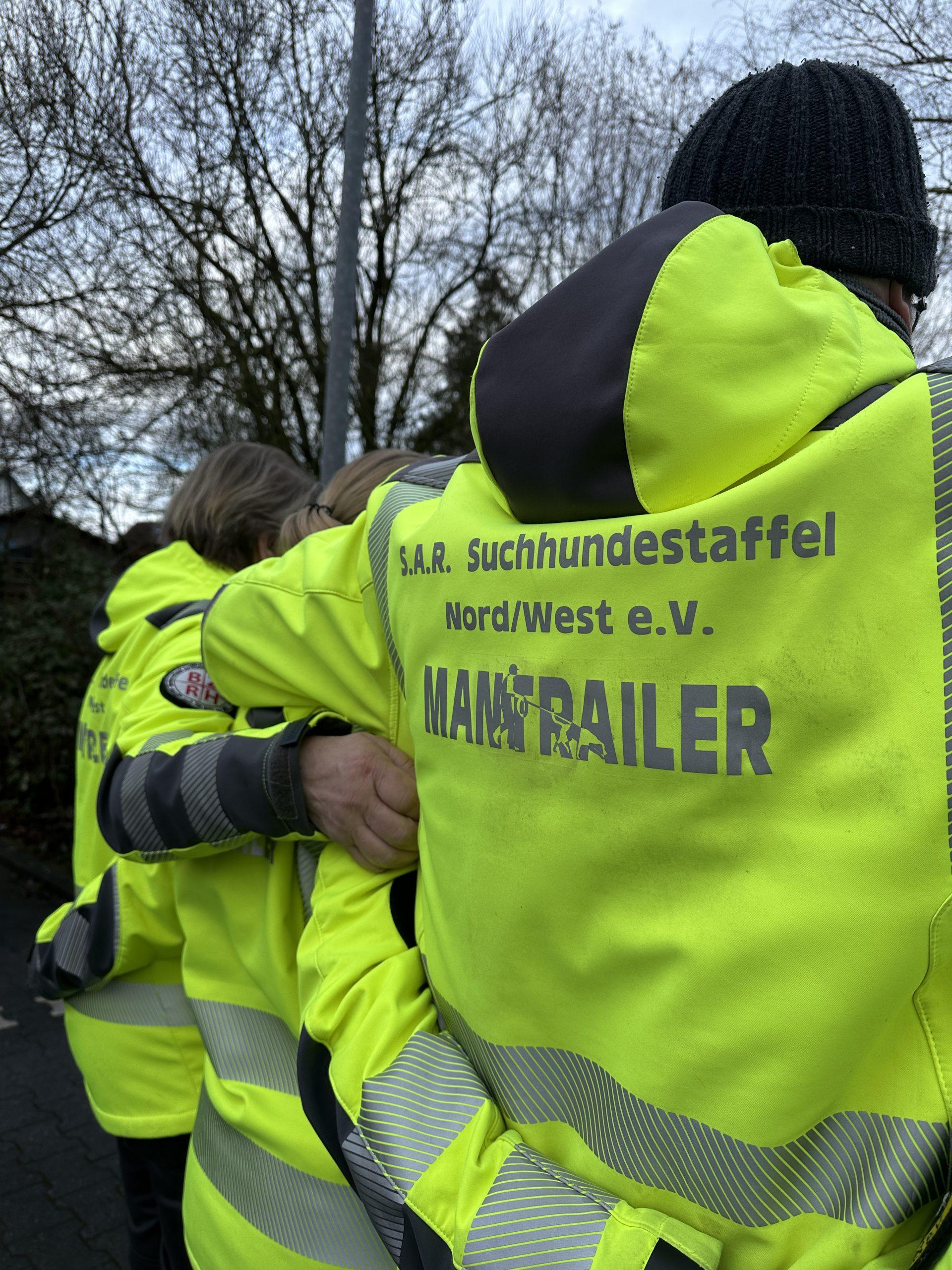 Vier Mitglieder der S.A.R. Suchhundestaffel Nord/West e.V. in gelber Einsatzkleidung mit der Aufschrift ‚MANTRAILER‘ stehen Schulter an Schulter, die Arme umeinander gelegt, Rücken zur Kamera.