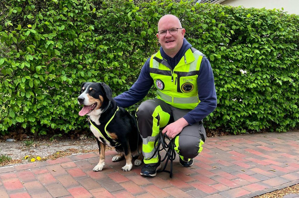 Mitglied der S.A.R. Suchhundestaffel Nordwest in Einsatzkleidung hockt neben seinem Suchhund, beide lächeln entspannt in die Kamera.