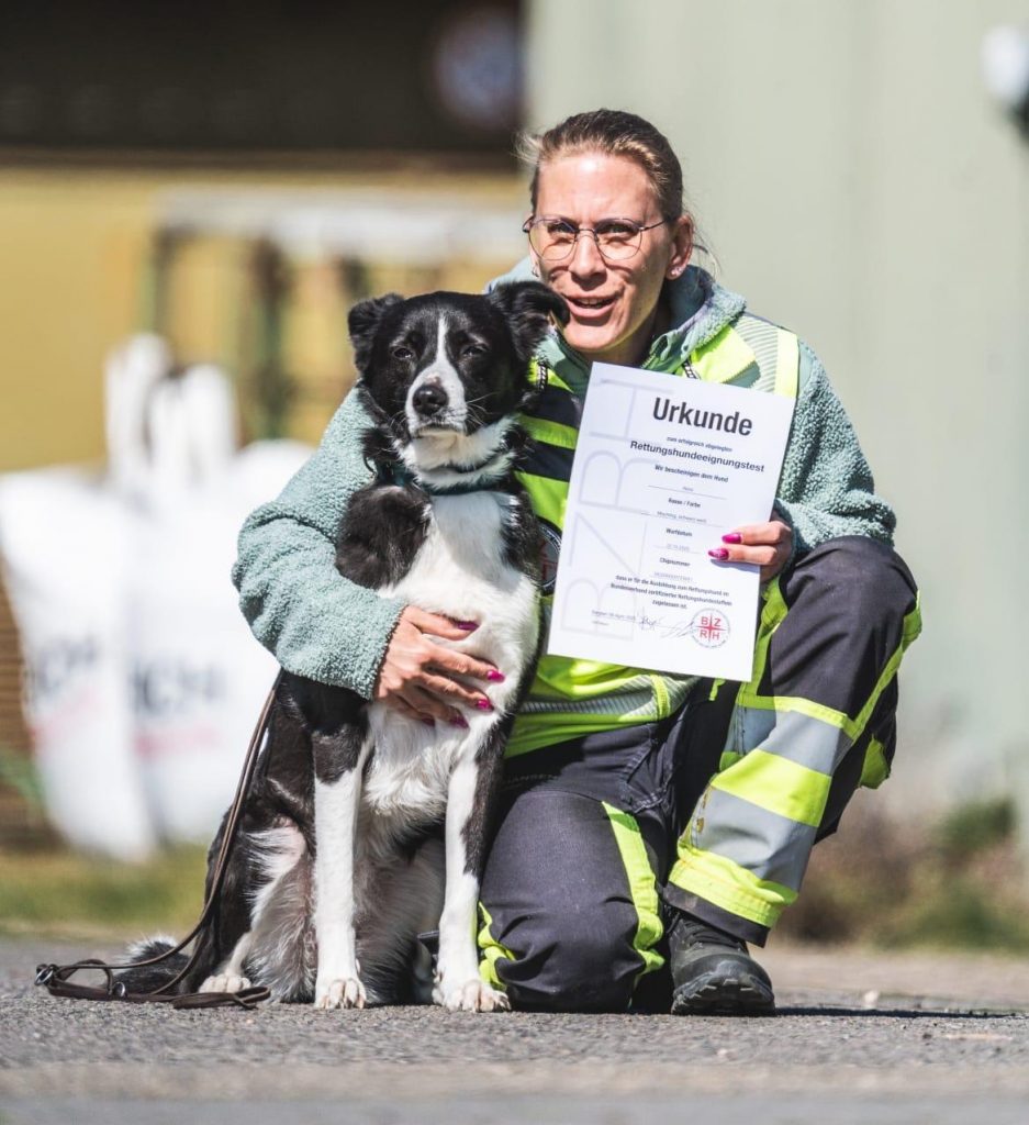 Hundeführerin der S.A.R. Suchhundestaffel Nordwest kniet neben ihrem Rettungshund und hält eine Urkunde des bestandenen Prüfungsnachweises in der Hand.